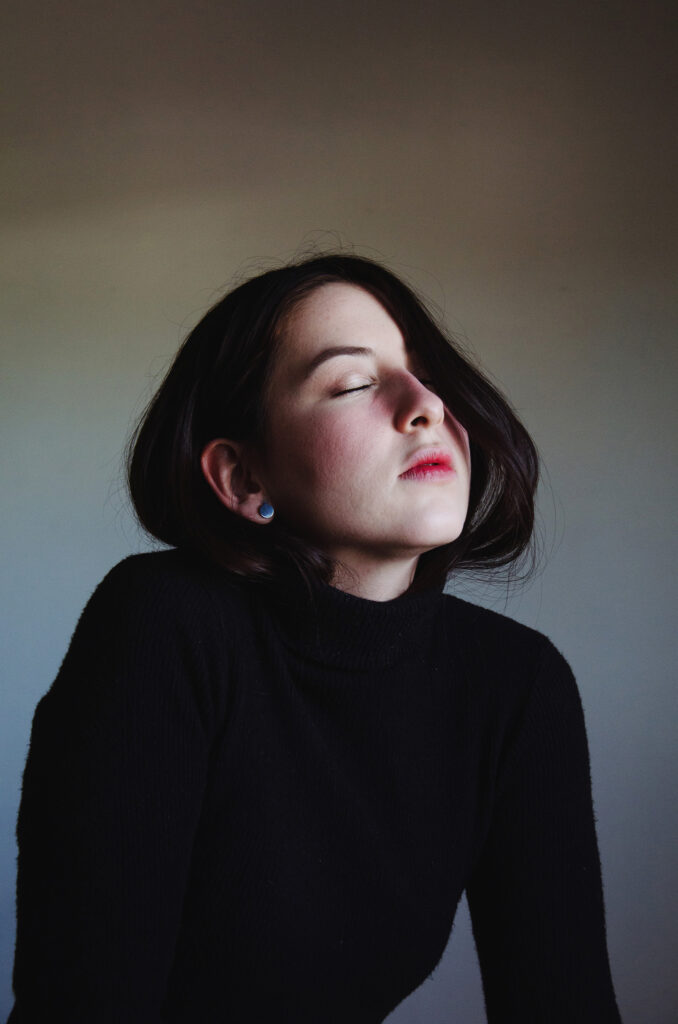 Gabriela Inzunza is captured in a serene moment with her eyes closed exuding a sense of calm. She wears a black turtleneck sweater and has a blue earring in her left ear. The background is a gradient of muted colors and the lighting casts a soft glow on her face.